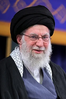 Ayatollah Ali Khamenei, Supreme Leader of Iran, wearing black robes and turban, seated in front of Iranian flag, portrait for biography and legacy article.
